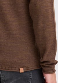 Brun stickad tröja med texturerad finish och ribbade ärmar. Har en liten läderetikett i fållen. Passform som är exakt och med släta linjer.