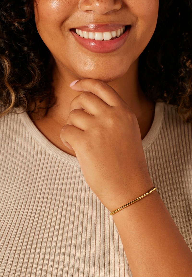 Gouden armband met een kralenontwerp, rustend op een hand. De hand raakt zachtjes de kin aan, tegen een beige top met een textuur. Soepele textuur.