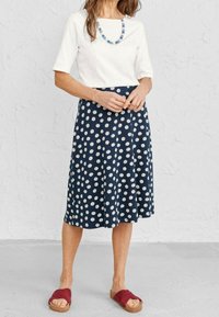 Haut blanc à manches courtes associé à une jupe marine à pois blancs. Le mannequin porte des sandales maroon au design torsadé.