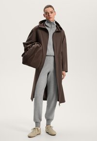 Brown long coat with a hood, gray zip-up top, gray sweatpants, and beige shoes. Model holding a matching brown bag. Smooth textures.