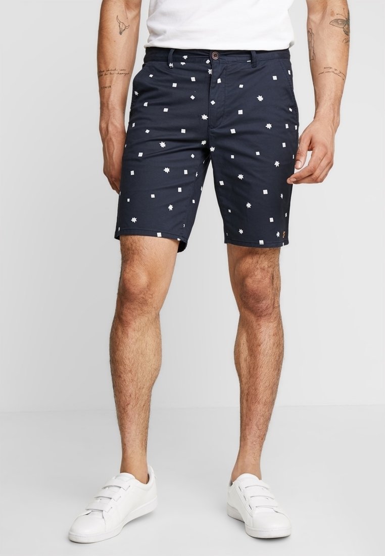 Shorts bleu marine avec des motifs géométriques blancs, fabriqués en tissu léger, avec une fermeture à bouton et des poches latérales. Baskets blanches.