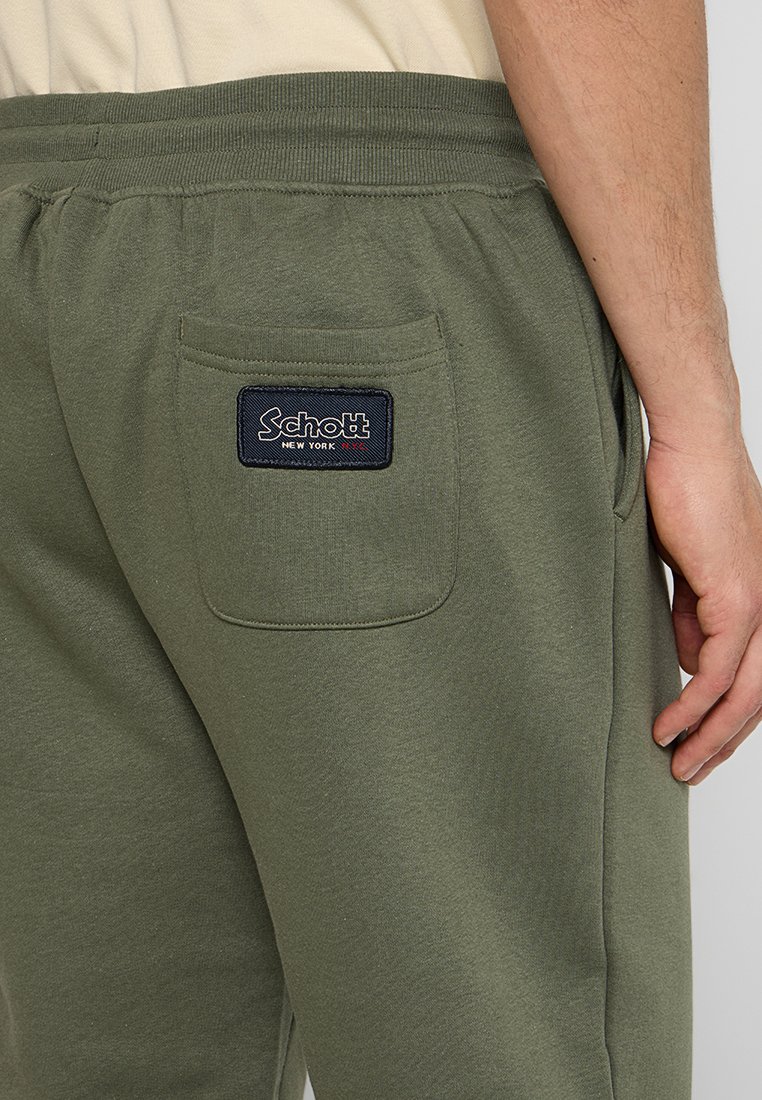 I pantaloni della tuta verde oliva presentano una tasca posteriore con un'etichetta Schott New York blu navy. Il materiale appare morbido e ha una consistenza liscia.