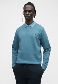 Blå poloskjorte med lange ermer, krage og ribbestrikkede mansjetter. Har en liten gul brodert logo på venstre bryst.