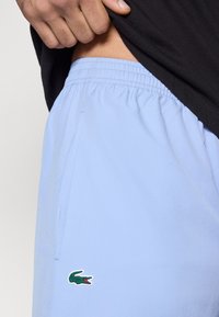 Ανοιχτό μπλε σορτς από διαπνέον ύφασμα, με ελαστική μέση και πλαϊνή τσέπη, με πράσινο λογότυπο κροκόδειλου στο αριστερό πόδι.