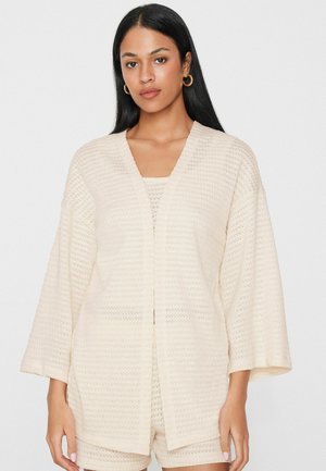 Frau mit langen schwarzen Haaren trägt einen cremefarbenen, strukturierten Cardigan mit offenem Vorderteil und passende Shorts, verziert mit goldenen Creolen.