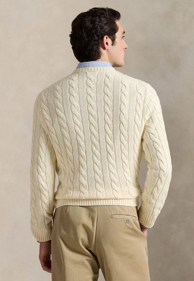 Homme portant un pull crème en maille torsadée sur une chemise bleu clair à col et un pantalon beige, de dos sur un fond neutre uni.