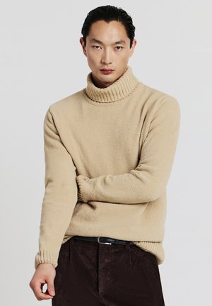 Beige Rollkragenpullover mit geripptem Kragen und Saum, der ein strukturiertes Strickmuster zeigt. Getragen mit dunklen Cordhosen.
