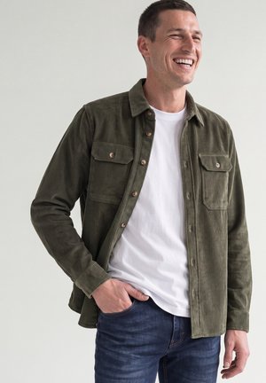 Mann lächelt, trägt ein offenes dunkelgrünes Cordhemd über einem weißen T-Shirt mit einer Hand in der Tasche der blauen Jeans vor hellem Hintergrund.