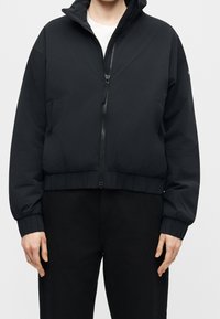 Personne portant une veste noire zippée avec poignets et ourlet élastiques, associée à un pantalon noir et une chemise blanche en dessous.