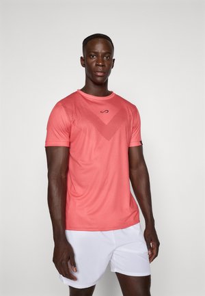 Athletic man wearing coral short-sleeve shirt with dotted chevron pattern and white shorts, standing against plain light background.