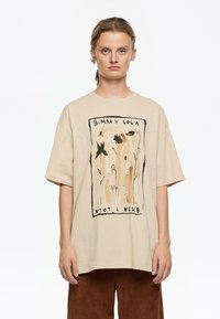 Beige suurenev t-särk musta ja pruuni lilleillustratsiooniga, mis on raamitud mustast kontuurist. Lühikesed varrukad ja ümar kaelus.