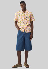 Camisa rosa de manga corta con estampado de limones y limas, combinada con pantalones cortos de denim azul sueltos y sandalias marrones. Tejido suave, corte relajado.