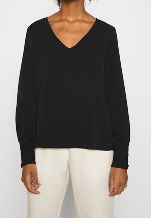 Vrouw die een zwarte blouse met lange mouwen, V-hals en geknoopte manchetten draagt, samen met ecru broek, staande tegen een effen achtergrond.