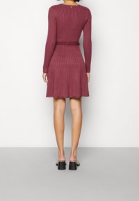 Robe tricotée bordeaux avec texture côtelée, manches longues et jupe évasée. Présente une taille ajustée et un col rond avec un détail de bouton.