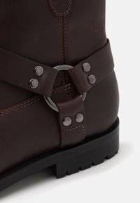 Brown leather boot featuring a silver metal ring and two studs on a leather strap, with contrasting stitching and a black rubber sole.
