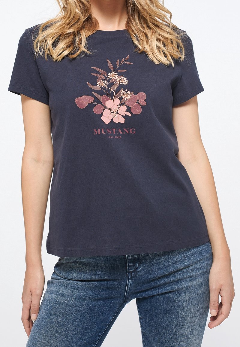 Kvinna iklädd en marinblå t-shirt med rosa blommig grafik och texten "MUSTANG est. 1932", tillsammans med blåjeans.