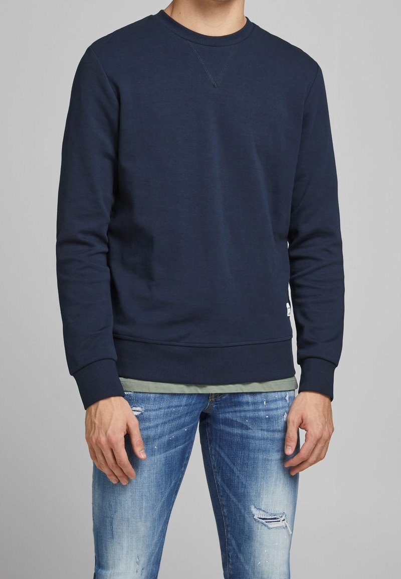 Mörkblå sweatshirt med rund halsringning, ribbade mudd och fåll, samt lätt textur. Bärs över en grön skjorta, i kombination med slitna jeans.