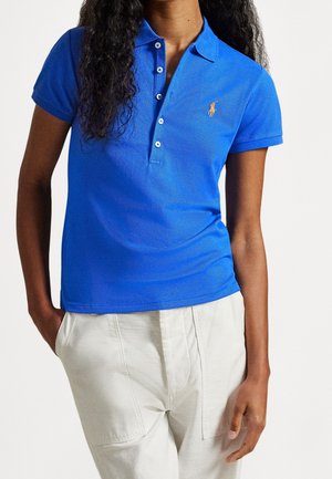Mujer con una camiseta tipo polo de manga corta de color azul brillante con un pequeño logo amarillo, combinada con pantalones de color claro.