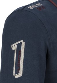 Tmavě modrá bavlněná košile s kulatým výstřihem, obsahující strukturované vyšívané číslo "1" v béžové barvě a červených akcentů, a vyšívaný text na rameni.