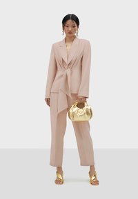 Svijetlo ružičasti blejzer s vezanjem u struku, odgovarajuće hlače, zlatna metalna torba i zlatne sandale s potpeticom. Glatka tkanina s krojem koji lijepo pristaje.