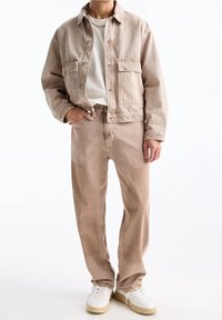 Beige denimjakke med to forlommer, parret med matchende lyse bukser. Hvid t-shirt under. Model iført hvide sneakers.