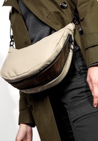 Beige Nylon Umhängetasche mit geschwungener Form, ausgestattet mit einem braunen unteren Abschnitt und einem oberen Reißverschluss. Verstellbarer Trageriemen und sichtbare Hardware-Details.