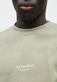 Olivzaļš džemperis no mīksta kokvilnas ar apaļu kaklu un baltu uzrakstu "JJ Studios" uz krūškurvja.