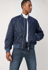 Marinejakke med en jevn, blank overflate, ribbede mansjetter og midjebånd. Har glidelås foran og sidelommer. Stylet med en hvit skjorte.