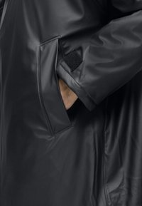 Îmbrăcăminte exterioară neagră, realizată dintr-un material din vinil neted și lucios. Include o buzunare lateral și un accent cu arici pe mânică pentru închidere.