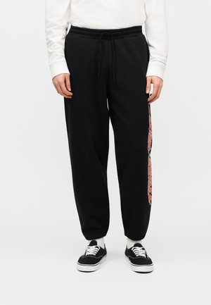 Άτομο που φοράει μαύρο παντελόνι φόρμας με κόκκινη και λευκή ρίγα με λεοπάρ σχέδιο στο πλάι, μαύρα αθλητικά παπούτσια, λευκές κάλτσες και λευκό μακρυμάνικο πάνω μέρος.