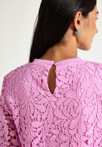 Nainen, jolla on pitkät tummat hiukset, käyttää kimaltavia sydämenmuotoisia korviksia ja vaaleanpunaista pitsipaitaa, jossa on avaus niskan takana.