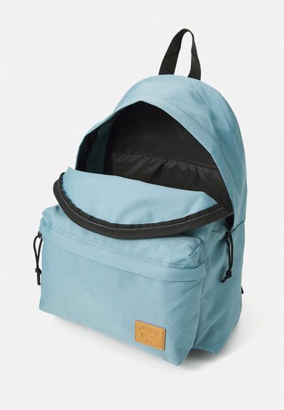 Sac à dos bleu clair avec une poche zippée à l'avant, doublure noire et un patch en cuir avec logo. Il dispose de bretelles ajustables et d'une poignée sur le dessus.