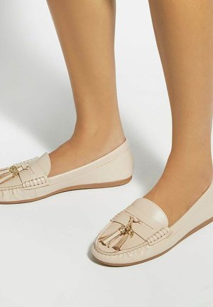 Feet wearing beige leather loafers with tassels, shown against a plain light background.