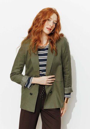 Femme aux cheveux roux portant un blazer vert olive sur une chemise à rayures et un pantalon foncé, avec un collier à chaîne dorée et des bracelets, debout devant un fond blanc.
