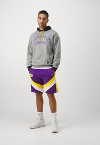 Sudadera gris con gráfico en zigzag morado, cordón amarillo y bolsillo; pantalones cortos morados con rayas blancas y amarillas; zapatillas blancas.