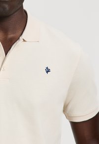 Világos bézs színű pólóing, texturált anyagból készült. A mellkason sötétkék hímzett korall logó található, és klasszikus galléros kialakítás jellemzi.