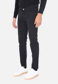 Uomo che indossa pantaloni neri slim-fit, maglione nero e sneakers bianche basse con suola beige, in piedi su uno sfondo bianco.