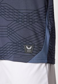 Camisola desportiva azul-marinho com acabamento texturizado, padrão abstrato preto e uma etiqueta tecida com logótipo no lado inferior esquerdo.