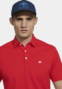 Rode polo shirt van textuurstof met een drieknops sluiting, voorzien van een wit logo op de linkerborst en een marineblauwe pet met een logo patch.