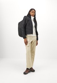 Giacca a vento nera, maglietta bianca con colletto alto, pantaloni cargo beige e scarpe da trekking marroni. Zaino nero con spallacci regolabili.