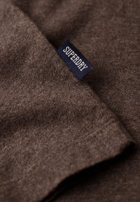 Brunt sweaterstof med en glat tekstur, prydet med et mørkeblåt mærke med "SUPERDRY" i hvid skrift. Nærbillede af vævningen.