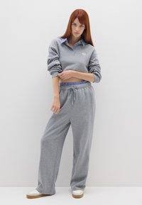 Sudadera gris con un collar a rayas y un logotipo, combinada con pantalones de chándal grises. Presenta un corte relajado y una textura de tela suave. Zapatillas blancas con suelas marrones.