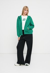 Sudadera verde con cremallera y dos bolsillos frontales, llevada sobre una camiseta blanca con el número 23. Combinada con pantalones anchos negros y zapatillas blancas.