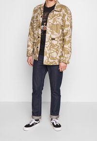 Uomo che indossa una giacca mimetica beige e bianca, una t-shirt nera con grafica, jeans scuri arrotolati e sneakers bianco e nere, in piedi su un pavimento chiaro.