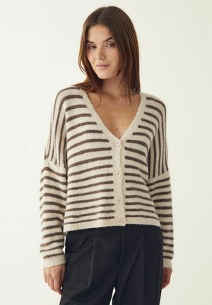 Naine, kes kannab beeži ja pruuni triibulist nööpidega kardigani pikkade varrukatega ning musti pükse, seisab ühetoonilise heleda tausta ees.