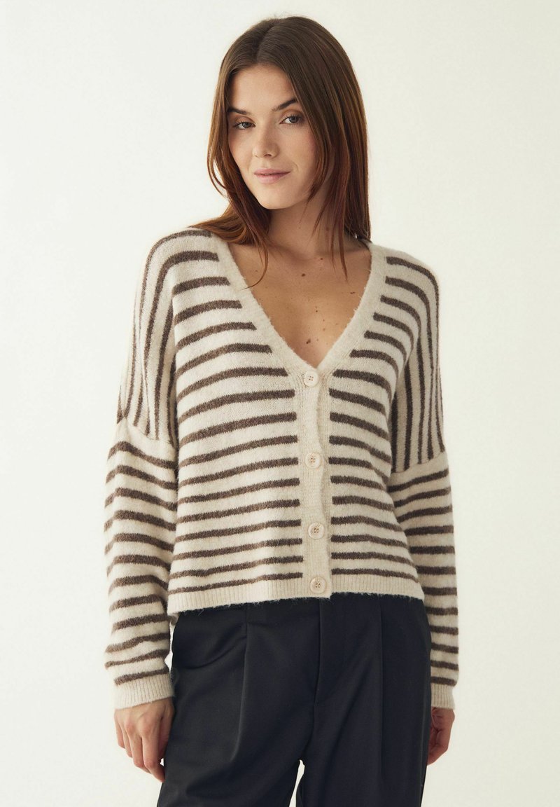 Vrouw die een beige en bruin gestreepte gebreide vest met knopen en lange mouwen draagt, gecombineerd met een zwarte broek, staand tegen een effen lichte achtergrond.