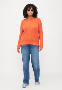 Oranger, gerippter Strickpullover mit langen Ärmeln, kombiniert mit hellblauen, geraden Jeans und grauen Turnschuhen, die ein Logo-Detail aufweisen.