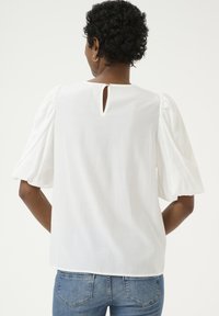 Blusa bianca con maniche corte a palloncino, aperture a chiave sul retro, texture morbida e vestibilità ampia; abbinata a jeans in denim, senza motivi visibili.