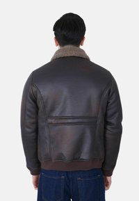 Uomo che indossa una giacca di pelle marrone scuro con un colletto in shearling e jeans blu, girato di spalle su uno sfondo bianco semplice.