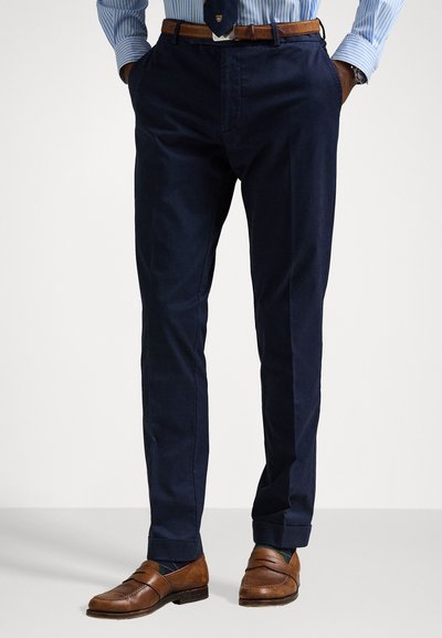 Marineblaue maßgeschneiderte Hosen mit glatter Textur, klassischem Schnitt und klaren Falten. Kombiniert mit braunen Slippern und einem gestreiften Hemd.